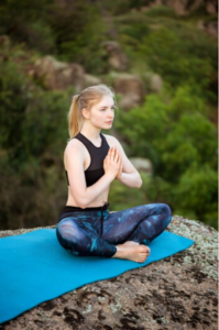 A woman in athletic wear meditating peacefully on a rock, hands in Namaste, with a tranquil forest in the background