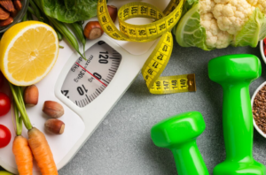 A bathroom scale with a measuring tape, green dumbbells, and a selection of healthy foods like vegetables and nuts on a grey background, symbolizing the components of a weight loss journey