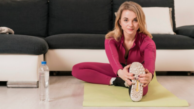 A woman in a pink tracksuit stretching on a yellow mat in her living room, with a bottle of water beside her.