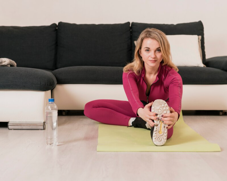 A woman in a pink tracksuit stretching on a yellow mat in her living room, with a bottle of water beside her.