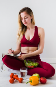 A woman in red sportswear sitting cross-legged, planning a diet with fresh vegetables and dumbbells beside her